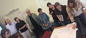 Acht Frauen und Männer stehen an einem Tisch und blicken auf ein Zettel, die darauf liegen.