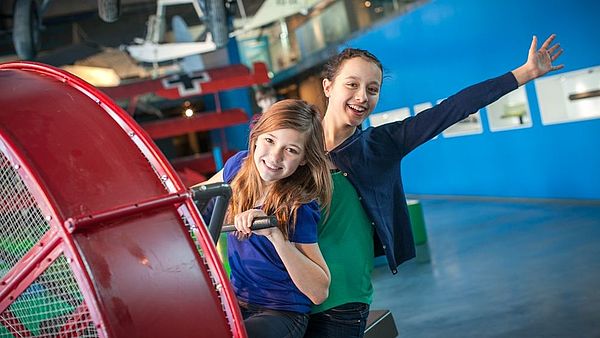 Zwei Jugendliche sitzen an einem roten Exponat der Ausstellung. Ein Mädchen hält sich daran fest. Das andere streckt lachend einen Arm in die Luft.