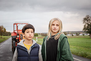 Iran Selcuk und Lotte Engels stehen auf einer Straße. Hinter ihnen steht ein roter Traktor. Links und rechts ist Wiese. Im Hintergrund befindet sich ein Ort.