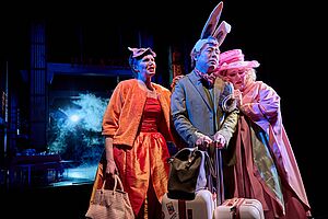 3 Menschen in Kostümen stehen auf einer Bühne. Sie stehen nah beieinander.