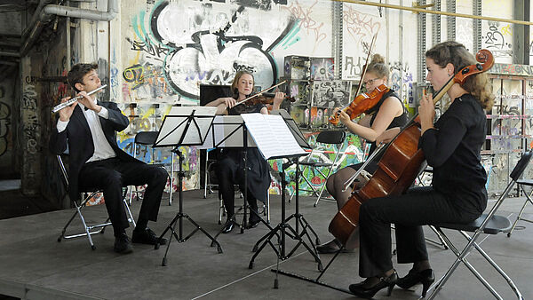 Ein musikalisches Quartett sitzt vor einer Graffiti besprühten Wand und spielt Querflöte, Geige und Cello. 