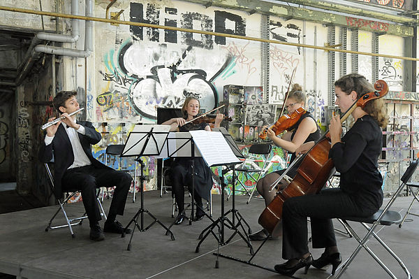 Ein musikalisches Quartett sitzt vor einer Graffiti besprühten Wand und spielt Querflöte, Geige und Cello. 