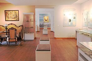 Einer der Museumsräume. In der Mitte stehen Bänke zum Verweilen. Im Raum befinden sich mehrere Glasvitrinen mit Ausstellungsstücken. Links stehen ein Sofa, Stühle und ein Tisch. An den Wänden hängen Bilder. Ein Durchgang gibt den Blick frei auf nachfolgende Räume.