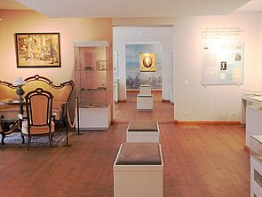 Einer der Museumsräume. In der Mitte stehen Bänke zum Verweilen. Im Raum befinden sich mehrere Glasvitrinen mit Ausstellungsstücken. Links stehen ein Sofa, Stühle und ein Tisch. An den Wänden hängen Bilder. Ein Durchgang gibt den Blick frei auf nachfolgende Räume.