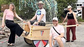 Vier Menschen stehen und sitzen in einem Holzwagen.