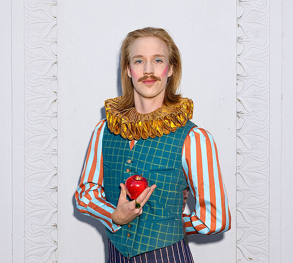 Ein Mann. Er spielt König Drosselbart und hält einen Apfel in der Hand.