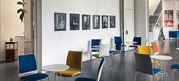 Ein Raum mit vielen Tischen und bunten Stühlen. An den Wänden hängen Aktfotografien von Klaus Ender.