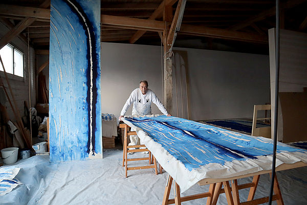 Günther Uecker steht in seinem großen Raum vor einem langen Tisch. Darauf liegt eine Stoffbahn mit blauem Muster. 