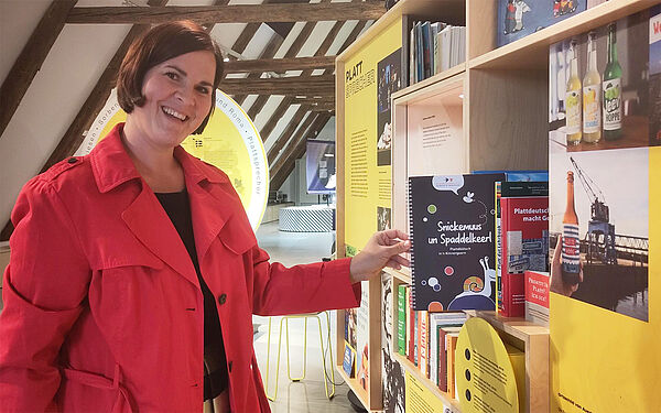 Eine Frau steht an einem Bücherregal. In dem Regal stehen viele Bücher. Sie zieht ein plattdeutsches Kinderbuch heraus.