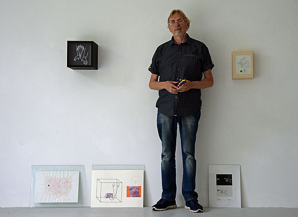 Udo Dettmann steht an einer Wand. Links und rechts neben ihm hängen Rahmen. Auf dem Boden lehnen Bilder an der Wand. 