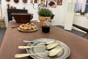 Auf einem Tisch steht ein Silberteller mit drei Silberlöffeln, eine Schale mit Nüssen, eine Flasche Essig, ein Kräutertopf und ein Abtropfsieb.