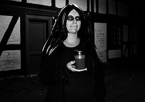 Eine blasse Person in dunklem Gewand steht im Dunkeln vor einem Fachwerkhaus.
