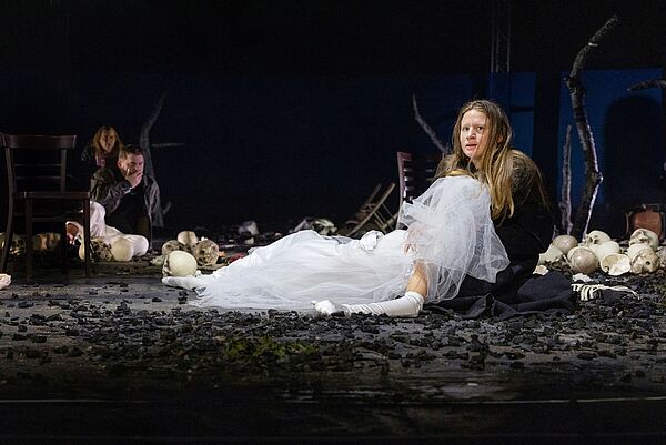 Eine Frau sitzt auf einer Bühne. Auf ihrem Schoß liegt eine Person in einem Brautkleid. Das Gesicht der Braut ist verschleiert. Neben ihnen liegen viele Totenschädel auf der Bühne. 
