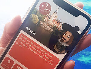 Eine Hand hält ein Handy. Auf dem Handy-Display ist die App für den Stadtrundgang geöffnet.