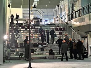 Auf einer Art Treppe verteilen sich einige Stühle. Im Raum stehen viele Leute.