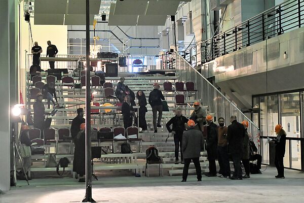 Auf einer Art Treppe verteilen sich einige Stühle. Im Raum stehen viele Leute.