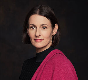Ein Porträt von Annett Renneberg. Sie trägt eine rötliche Jacke und steht vor einem schwarzen Hintergrund.