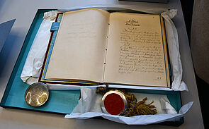 Eine geöffnete historische Handschrift liegt in einer türkisfarbenen Schutzbox. Neben dem Buch liegen ein Wachssiegel mit rotem Siegelstoff und Quaste sowie ein silbernes Siegelkapsel-Objekt.