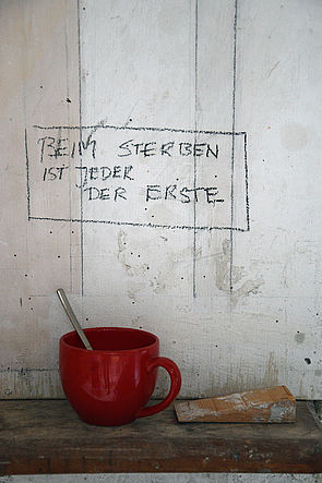 Auf einem Wandregal steht eine rote Tasse mit Löffel. An der Wand steht "Beim Sterben ist jeder der Erste." 