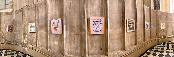 An einer Kirchenwand hängen nebeneinander sieben Bilderrahmen. Manche enthalten Bilder, andere geschriebene Worte.