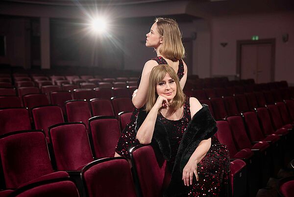 Zwei Schauspielerinnen. Sie sitzen Rücken an Rücken im Saal.