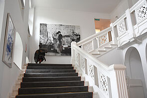 Eine Treppe mit weißen Geländer führt nach oben. Großformatige Bilder hängen an der Wand. Eine Tierskulptur steht auf der Treppe.