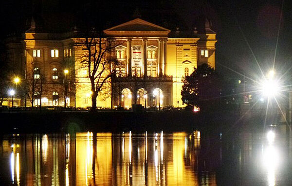 Außenaufnahme vom Theater in Schwerin. Es ist dunkel. Das Theater ist gelb angestrahlt und spiegelt sich im gegenüberliegenden Schweriner See.