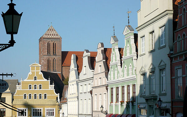 Alte Giebelhäuser stehen nebeneinander. Ihre Fassaden sind gelb, grün, grau und beige. Im Hintergrund ragt eine Kirche hervor.