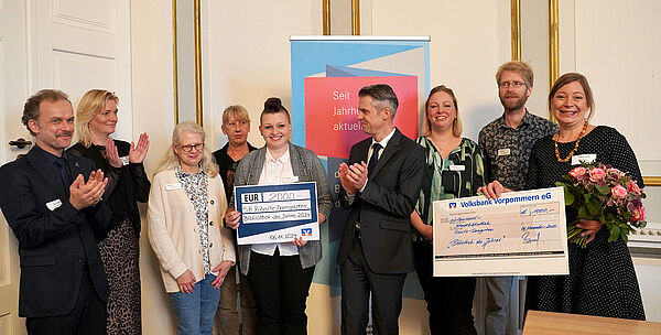 Neun Frauen und Männer stehen nebeneinander. Sie halten zwei große Geldschecks und Blumen in der Hand. Manche von ihnen klatschen.