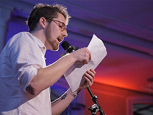 Diego Hagen Diego Hagen steht vor einem Mikrofon. Er hält eine Zettel in der Hand und spricht.