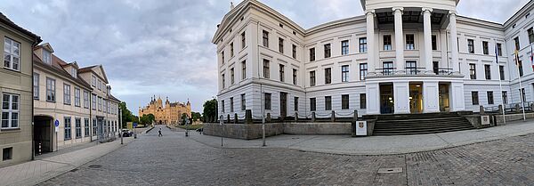 Rechts steht die Staatskanzlei. Sie hat eine weiße Fassade. Links befindet sich ein Fachwerkhaus. Zwischen beiden Gebäuden fällt die Sichtachse in der Ferne auf das Schloss. 