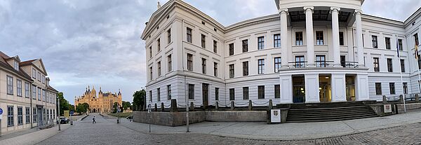 Rechts steht die Staatskanzlei. Sie hat eine weiße Fassade. Links befindet sich ein Fachwerkhaus. Zwischen beiden Gebäuden fällt die Sichtachse in der Ferne auf das Schloss.