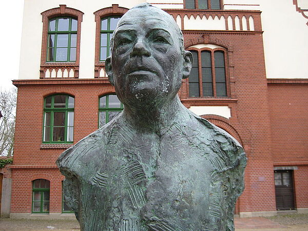 Eine Büste von Uwe Johnson. Dahinter steht ein Gebäude.
