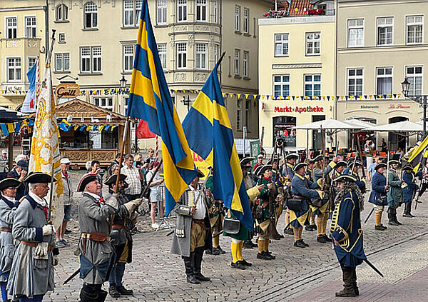 Viele Männer stehen auf dem Marktplatz in Wismar. Sie tragen Uniformen und Fahnen in den Farben blau und gelb. Sie sehen aus wie Soldaten.