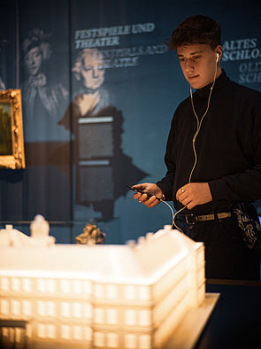 Ein junge steht mit Kopfhörern im Ohr und einer Fernbedienung in der Hand vor dem erleuchteten Modell des Schloss Neustrelitz.