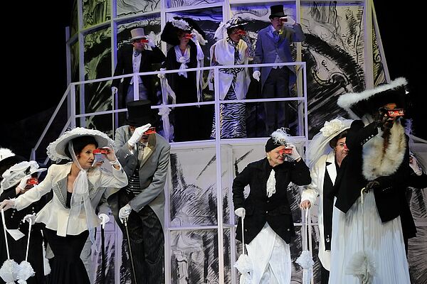 Zehn fein gekleidete Schauspielerinnen und Schauspieler stehen auf der Bühne und schauen durch Ferngläser. 