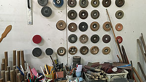 Ein Tisch voller Arbeitsuntensilien. Dazu gehören Pinsel, Schleifpapier und Kreide. An der Wand hängen runde Scheiben.