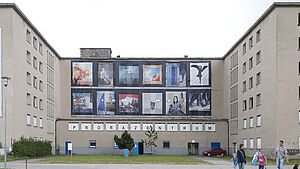 Block 5 auf Prora: Zwei leere Seitenblöcke, die mit einem Haus verbunden sind, an dem in Zweierreihe zwölf quadratische Plakate hängen. Weiter unten steht "Prorazentrum". Menschen gehen auf dem Gelände entlang.