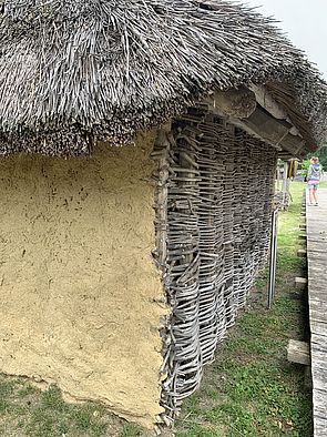 Eine Hütte mit Reetdach. Eine Wand besteht aus geflochtenen Weidenruten, eine andere ist mit Lehm verputzt.