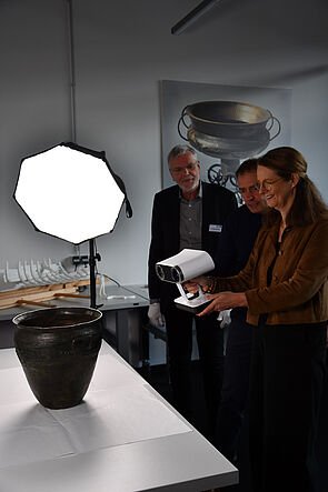 Drei Personen stehen in einem hellen Arbeitsraum vor einem Tisch. Eine Frau hält einen weißen 3D-Handscanner auf ein dunkles Tongefäß. Im Hintergrund ein großformatiges Foto eines bronzezeitlichen Kesselwagens.