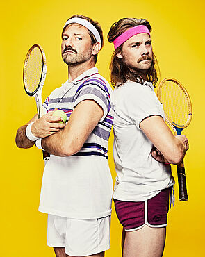 Zwei Männer stehen Rücken an Rücken. Sie tragen Tennisklamotten. Jeder hält einen Tennisschläger in der Hand.  
