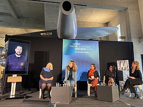 Auf einer Bühne sitzen fünf Frauen, darunter Ministerin Bettina Martin. Ein Mann ist der Runde digital über einen Bildschirm zugeschaltet.  