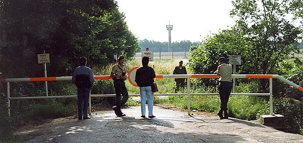 An der Grenze zwischen BRD und DDR. Vier Personen stehen auf der Westseite, darunter zwei Grenzschützer, ein Mann ist auf der anderen Seite zu sehen. Im Hintergrund: ein Grenzturm.