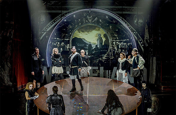 Eine Bühne. Auf der Bühne steht ein runder Tisch. Auf dem Tisch steht ein Mann. Um den Tisch herum stehen neuen Personen. 