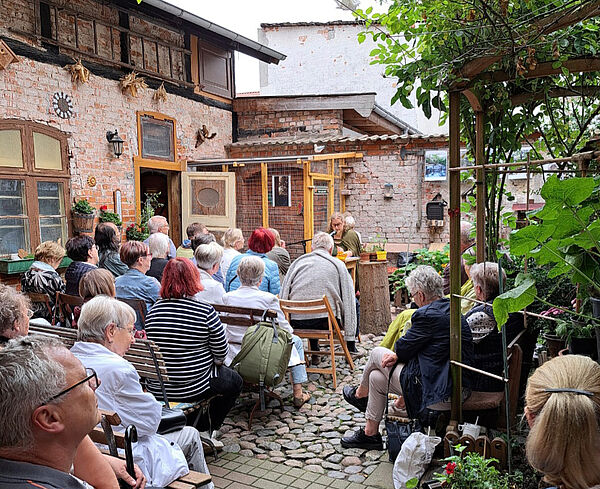 Ein Garten. Eine Frau liest aus einem Buch. Zuschauer hören zu.