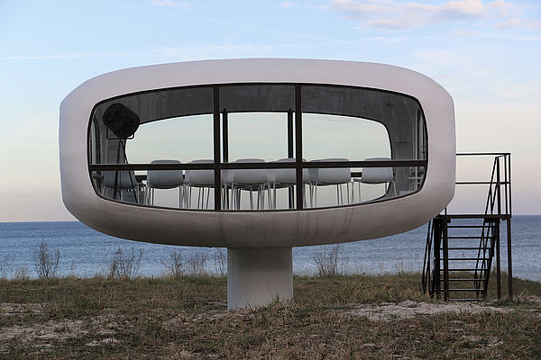 Ein weißer, ovaler Turm mit großen Glasfronten. Er steht auf einem Sockel. Im Raum stehen Stühle. Im Hintergrund ist die Ostsee. 