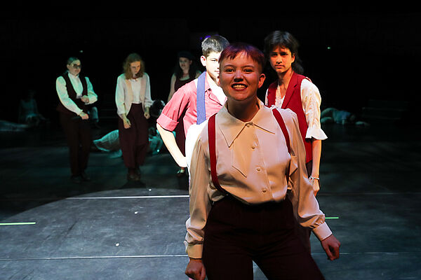 Eine Theaterbühne. Im Vordergrund steht eine junge Frau. Im Hintergrund stehen weitere Jugendliche.