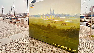 An der Promenade vom Museumshafen steht ein großes Plakat. Es zeigt die Silhouette von Greifswald, umgeben von Wiese, Bäumen und einigen Pferden. Auf dem Plakat steht auch die Internetadresse zum Jubiläum. Sie lautet www.caspardavid250.de. 
