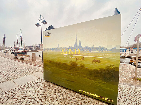 An der Promenade vom Museumshafen steht ein großes Plakat. Es zeigt die Silhouette von Greifswald, umgeben von Wiese, Bäumen und einigen Pferden. Auf dem Plakat steht auch die Internetadresse zum Jubiläum. Sie lautet www.caspardavid250.de.