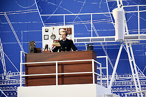 Ein Funker steht hinter einem Schreibtisch. Er liest einen Zettel. An der Wand hängen Uhren. Auf dem Tisch steht ein altes Telefon.